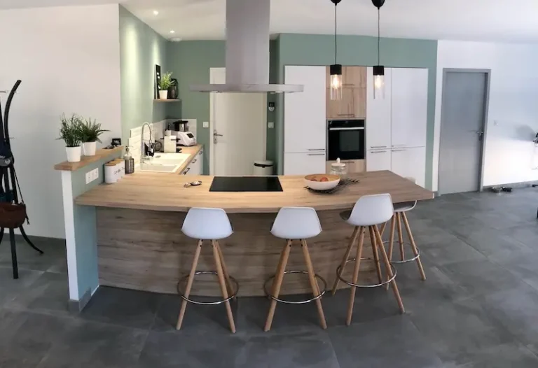 Vue d'ensemble d'une cuisine rénovée avec îlot central, four encastré, façades blanches sans poignées et sol carrelage grand format, par l'Entreprise Sarraud