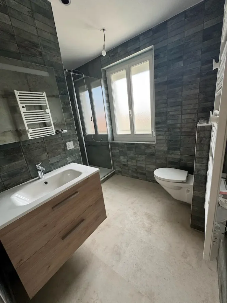 Vue complète d'une salle de bain rénovée à Coulon avec vasque sur meuble bois, WC suspendu et douche à l'italienne, ambiance minérale par l'Entreprise Sarraud