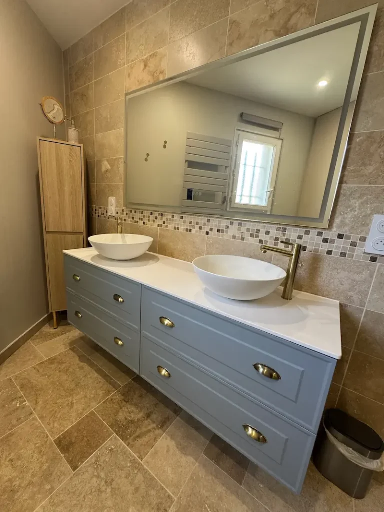 Salle de bain rénovée en travertin avec double vasque ronde blanche sur meuble bleu-gris à poignées laiton doré, robinetterie laiton brossé, par l'Entreprise Sarraud